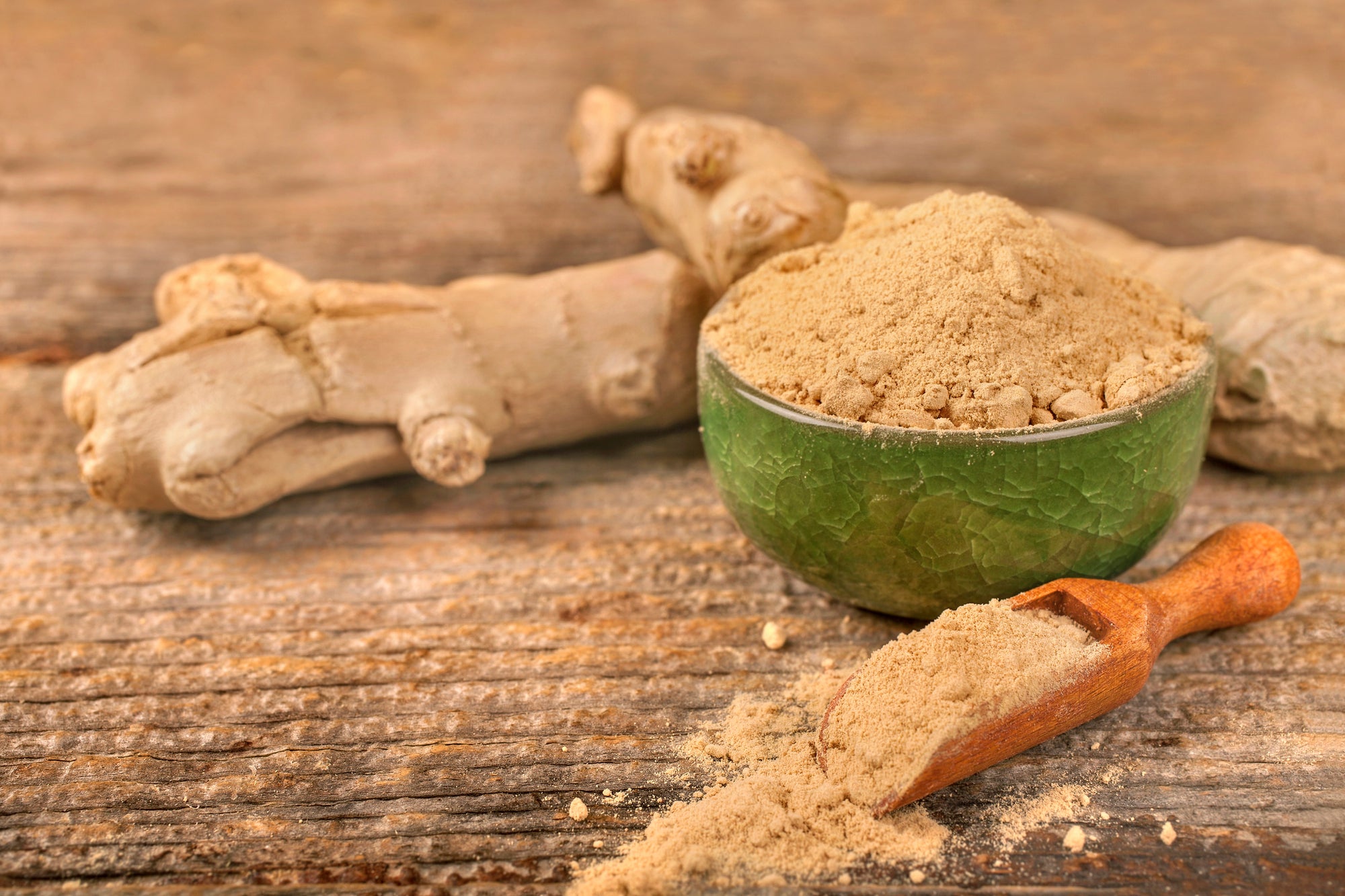 An image of ginger root powder in a bowl and wooden scoop and ginger root on a wooden table.